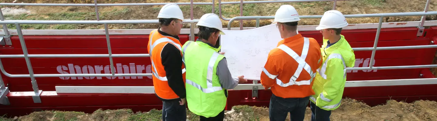 Four engineers, wearing their safety equipment, review blueprints for an excavation project while standing in front of a Steel Shoring Box equipped with an Edge Protection System, showcasing one of our Temporary Works Solutions services.