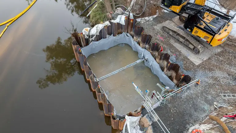 sheet pile cofferdam lined with protective sheeting contains water and sediment near an excavator on a riverbank construction site.