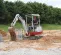 Red Takeuchi mini excavator being used to dig near a wooded area with a worker at the control
