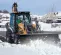 Orange and black Case 4 wheel drive extended cab backhoe loader using its bucket to push through heavy snow with a worker in the cab