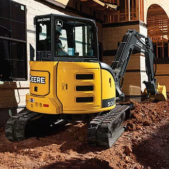 Yellow and black John Deere mini excavator beside a building at a construction site with a worker in the cab
