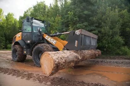 Orange and black Case wheel loader lifting a large tree trunk in the mud in a heavily wooded area