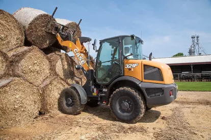 Orange and black Case wheel loader lifting bales of hay using a fork attachment with a worker in the cab