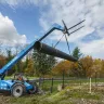 Blue Genie 8,000 lb. telehandler reach forklift with boom extended and holding pipe near a fence in a wooded area