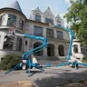 Two blue Genie 30-36 ft. towable extended boom lifts outside of a building