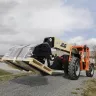 Montacargas de alcance con manipulador telescópico JLG naranja transportando un palé cargado con un trabajador en la cabina