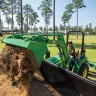 Green John Deere 30-39 HP 4WD compact backhoe loader dumping dirt into a container in a grassy area with a worker driving