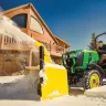 Green and yellow John Deere 30-39 HP 4WD compact backhoe loader blowing snow from in front of a home