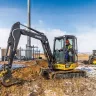 Yellow and black John Deere mini excavator digging dirt next to a chain link fence with a worker in the cab