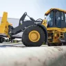 Yellow and black John Deere 4WD wheel loader moving pipe next to an 18-wheeler truck