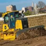 Yellow John Deere 110 HP bulldozer moving dirt next to a stone wall with a worker in the cab