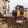 Yellow and black John Deere bulldozer with low ground pressure moving dirt beside a building with a worker in the cab