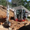 Red and white Takeuchi mini excavator being used to dig dirt and rocks near a house