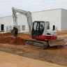 Red and white Takeuchi mini excavator being used to dig dirt next to a building