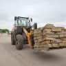 Orange and black Case wheel loader carrying a large pallet of stone blocks using a fork attachment with a worker in the cab