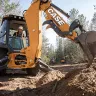 Orange and black Case 4 wheel drive backhoe loader using its boom to excavate dirt in a wooded area with a worker in the cab