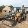 Orange and black Case 4 wheel drive extended cab backhoe loader at a construction site with dirt and rocks in its bucket and a worker in the cab