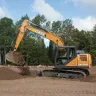 Orange and black Case excavator with boom lifting dirt in a wooded area and a worker in the cab