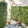 Orange Stihl hedge trimmer with 24 inch double-sided blade not in use and leaning against a hedge with personal protective equipment nearby