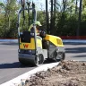 Yellow Wacker-Neuson ride-on double smooth-drum vibratory roller moving alongside a curb with a worker driving